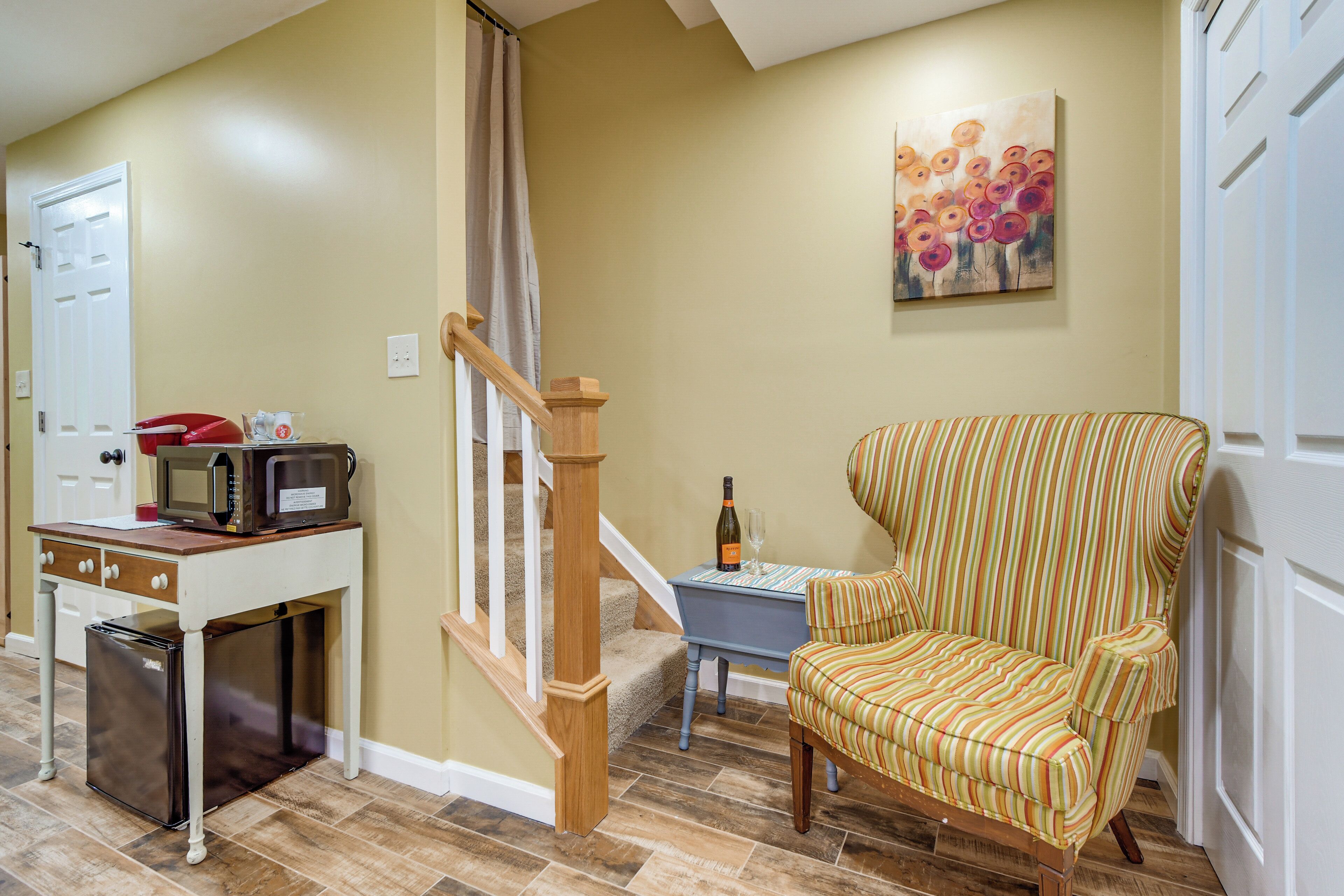 A living room with a striped armchair, a small table with a bottle and glass, a microwave, and a door.