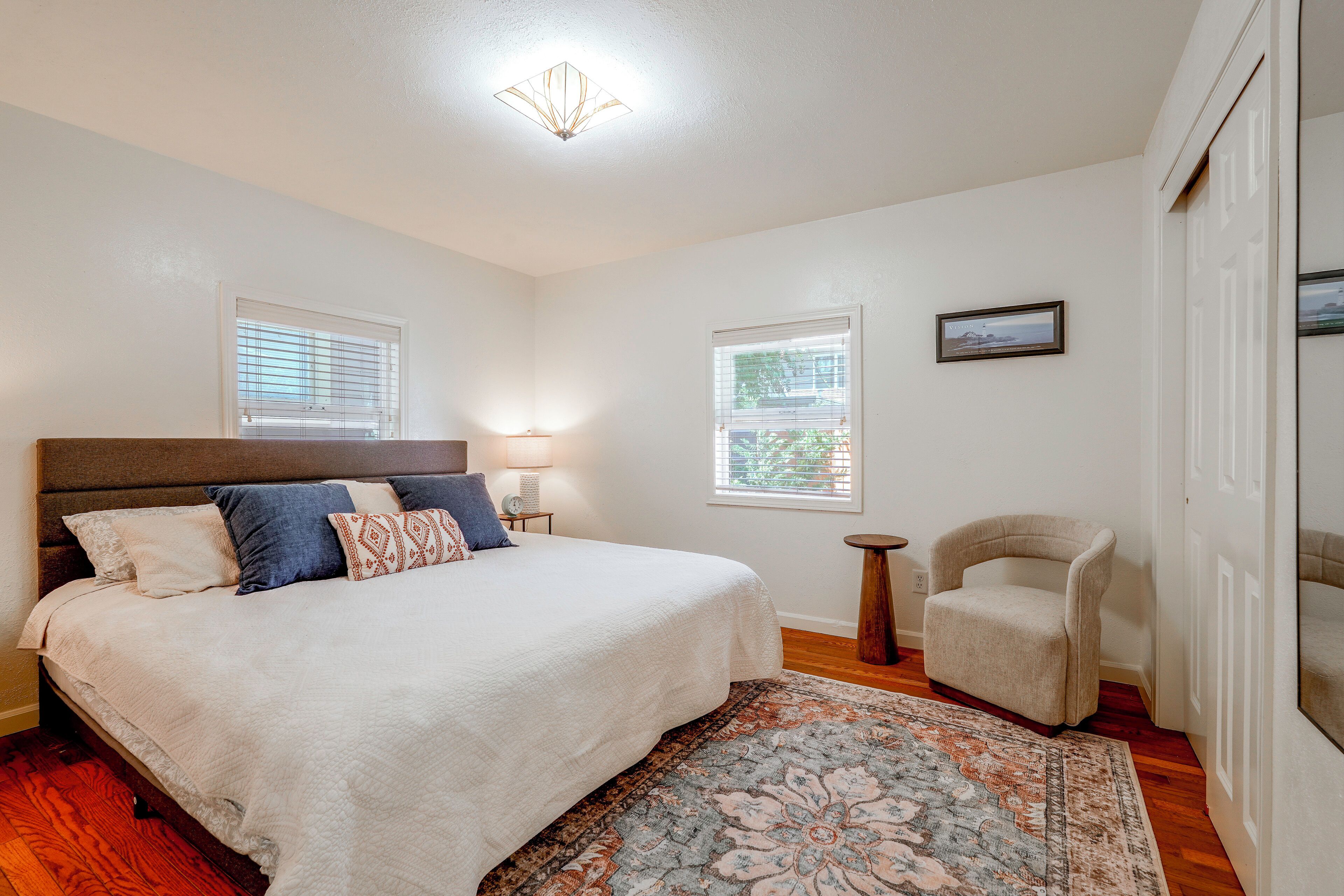 A bedroom with a bed, a chair, a rug, and two windows with blinds.