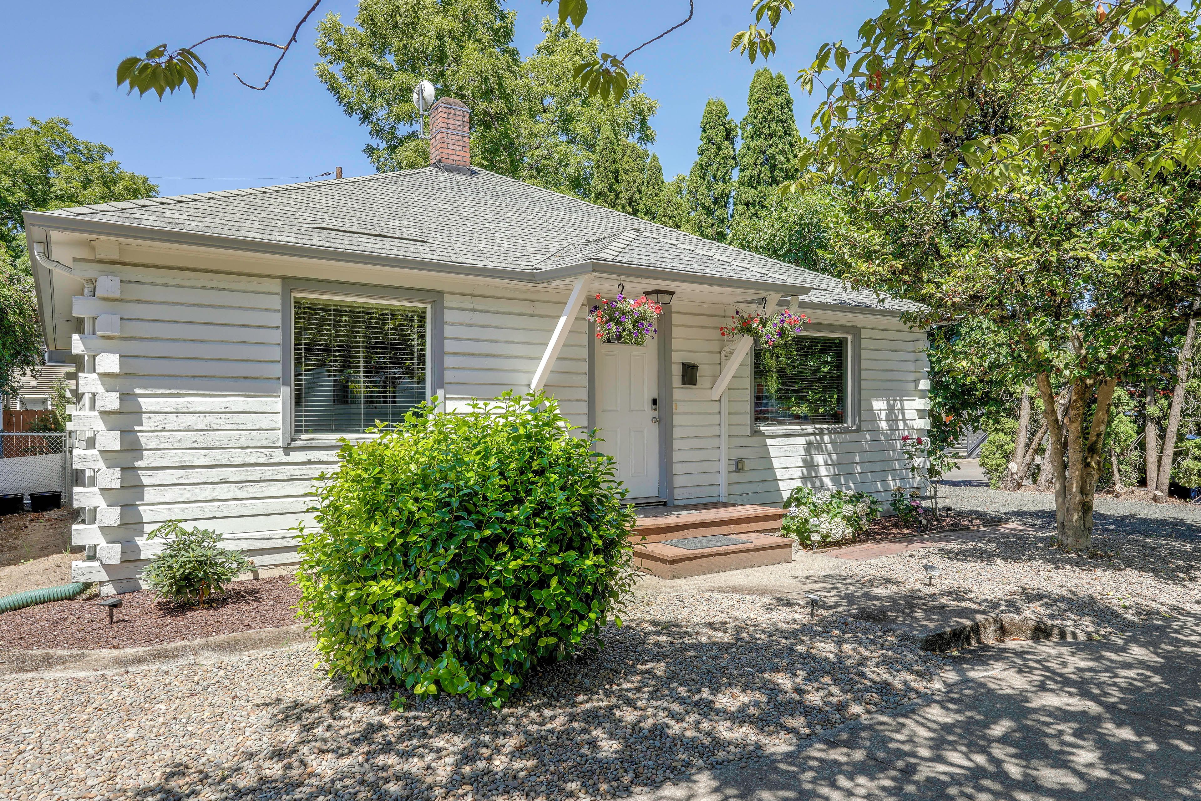 A small, single-story house with a front porch, a chimney, and a well-maintained garden with a hedge and gravel driveway.