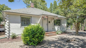 A small, single-story house with a front porch, a chimney, and a well-maintained garden with a hedge and gravel driveway.