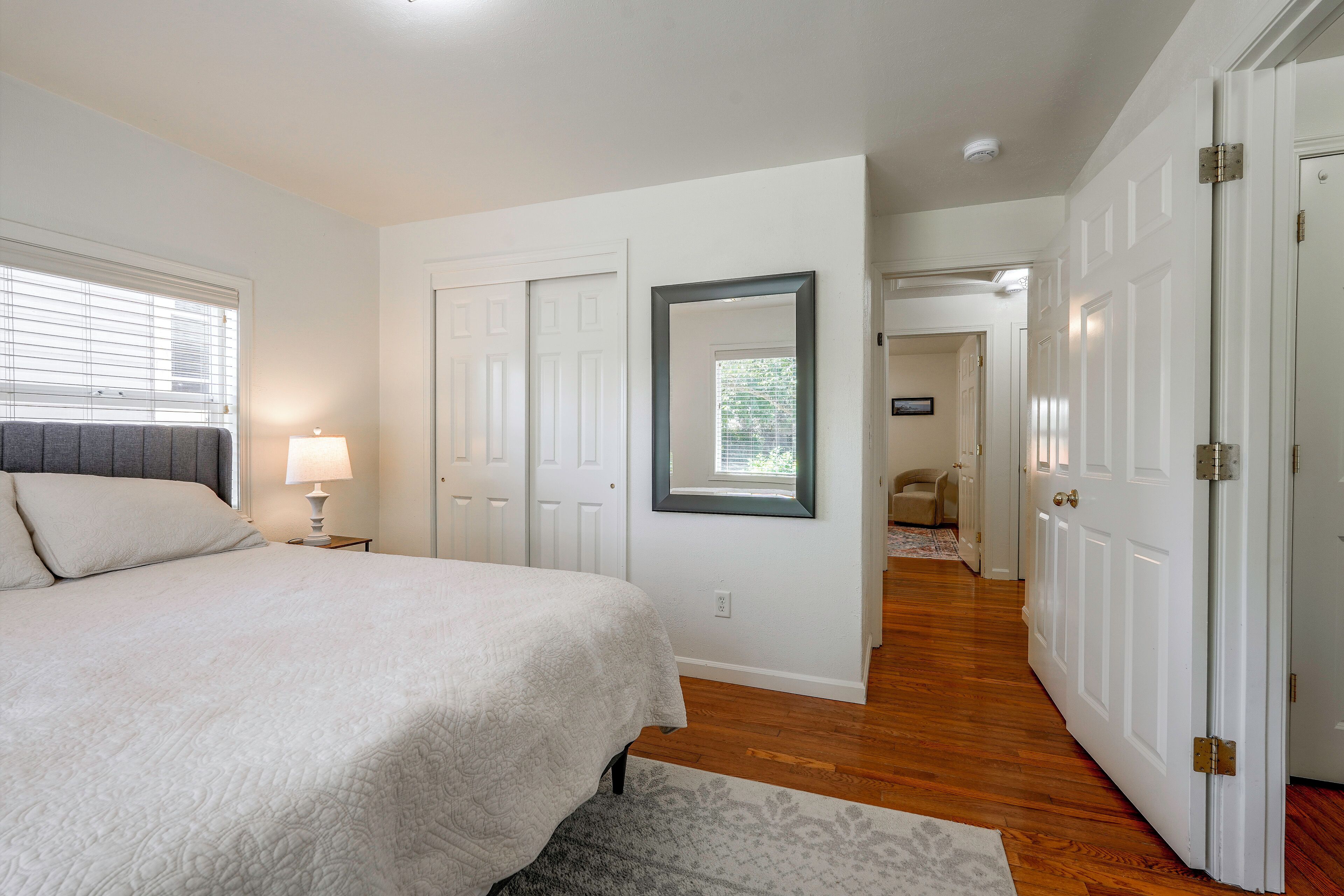 Un dormitorio con cama, mesita de noche, lámpara, espejo y una puerta que da a otra habitación.
