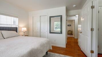 Un dormitorio con cama, mesita de noche, lámpara, espejo y una puerta que da a otra habitación.