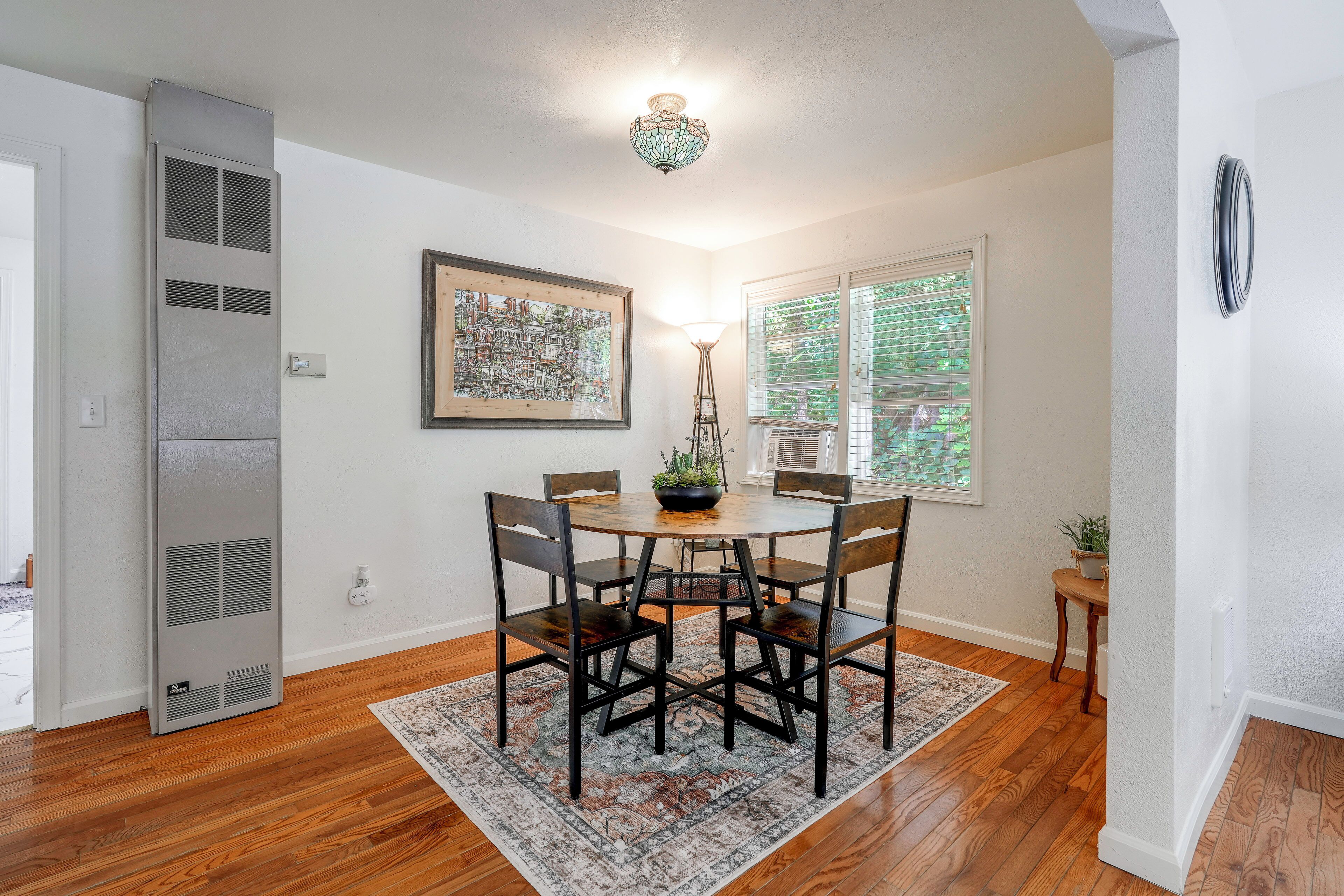 A dining area with a round table, four chairs, a patterned rug, a ceiling light fixture, and a framed artwork on the wall.
