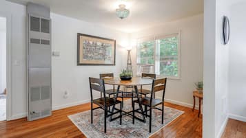 A dining area with a round table, four chairs, a patterned rug, a ceiling light fixture, and a framed artwork on the wall.