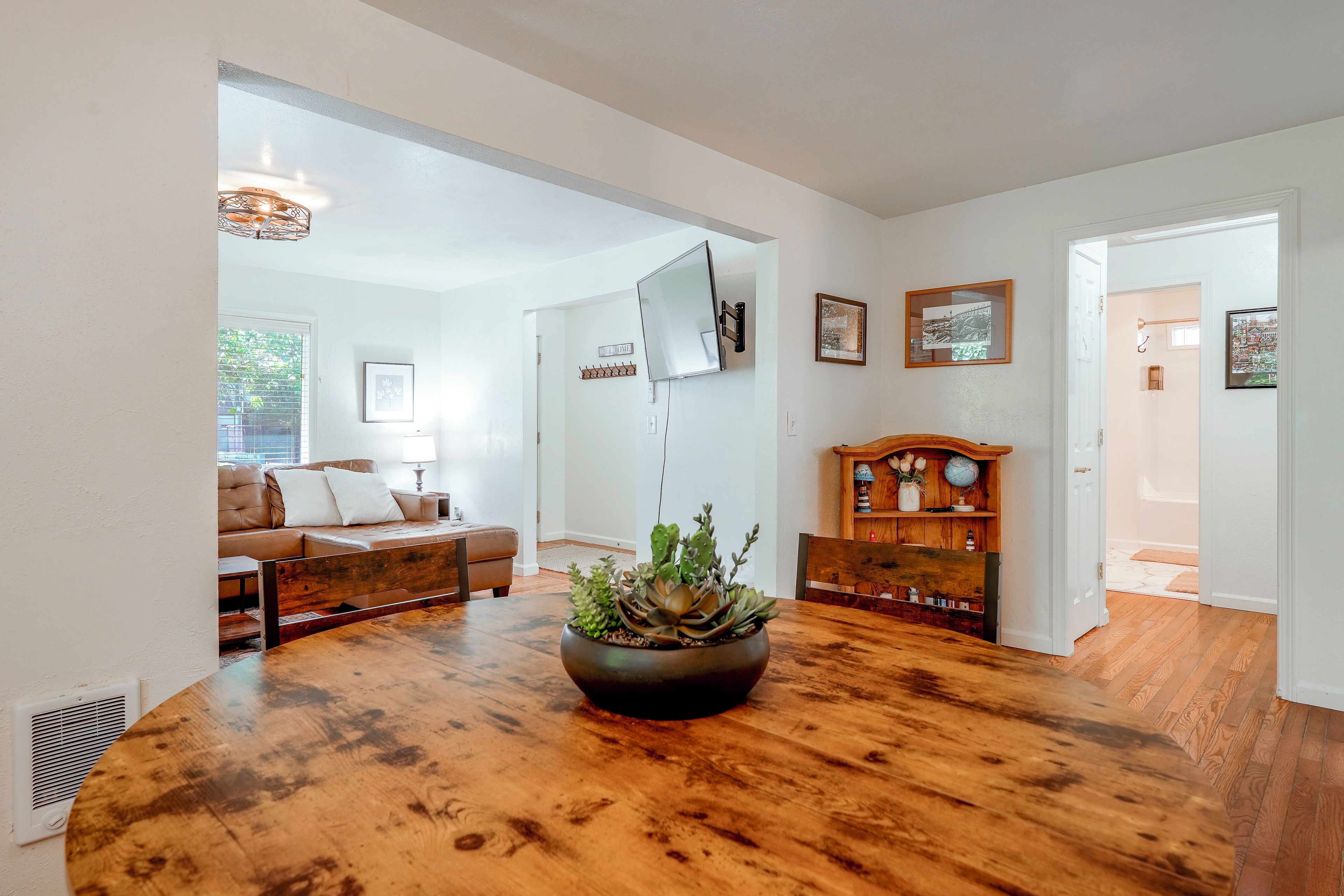 A spacious living room with a wooden floor, a leather sofa, a coffee table with a plant, and a wall-mounted TV.