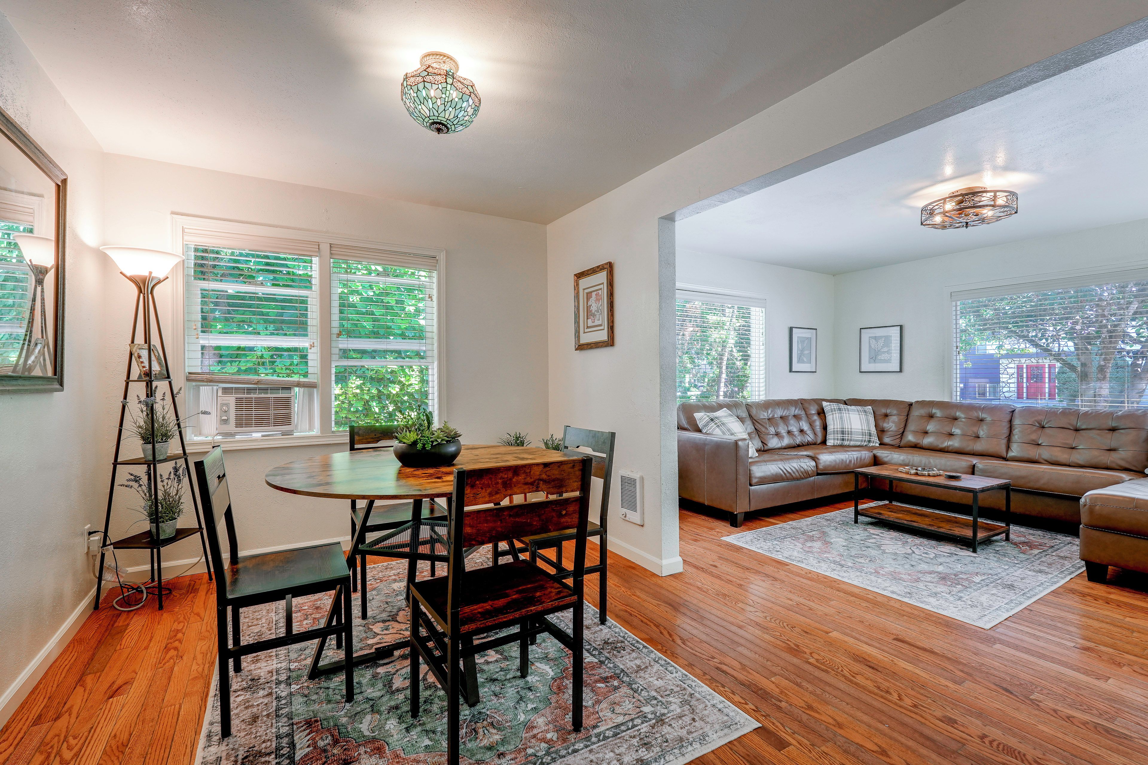 A spacious living area with a wooden floor, a dining table with chairs, a sofa, and a large window with blinds.