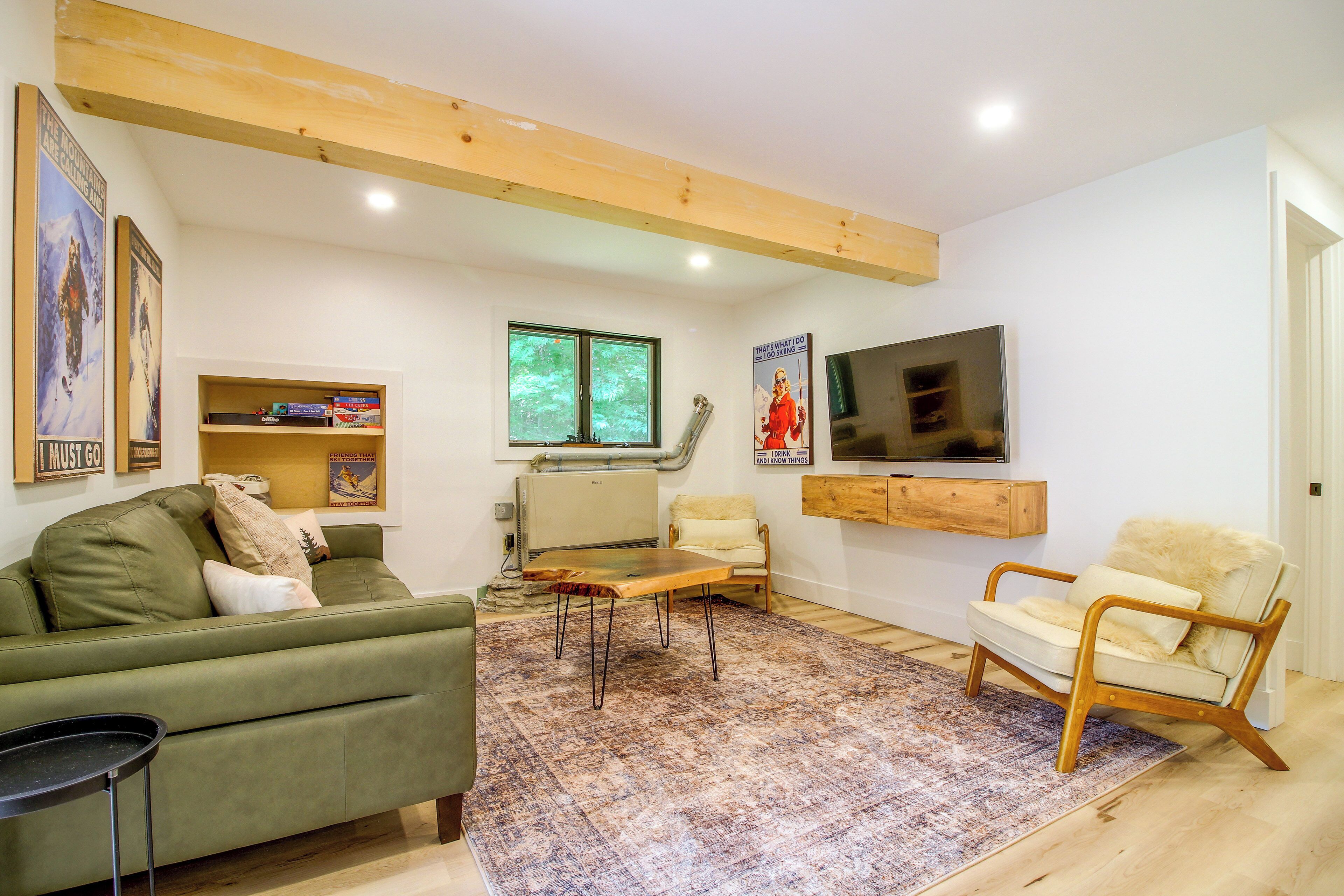 Una sala de estar con un sofá verde, una mesa de centro de madera, un tapete estampado, un televisor montado en la pared y una estantería con diversos objetos.