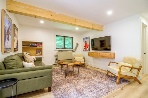 A living room with a green sofa, a wooden coffee table, a patterned rug, a television mounted on the wall, and a bookshelf with various items.
