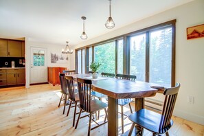 A dining area with a wooden table and chairs, large windows with a view of trees, and a kitchen with wooden cabinets.
