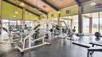 Uma academia com diversos equipamentos de exercício, grandes janelas e um ventilador de teto.