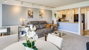A modern living room with a grey sofa, a coffee table, and a dining area with a white table. There are abstract paintings on the wall and a kitchen area in the background. - 1 Mi to Makaha Beach: 10th-floor Condo w/ Views (Waianae)