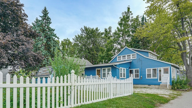 Une maison bleue avec une clôture blanche en bois, entourée de verdure et d’arbres.