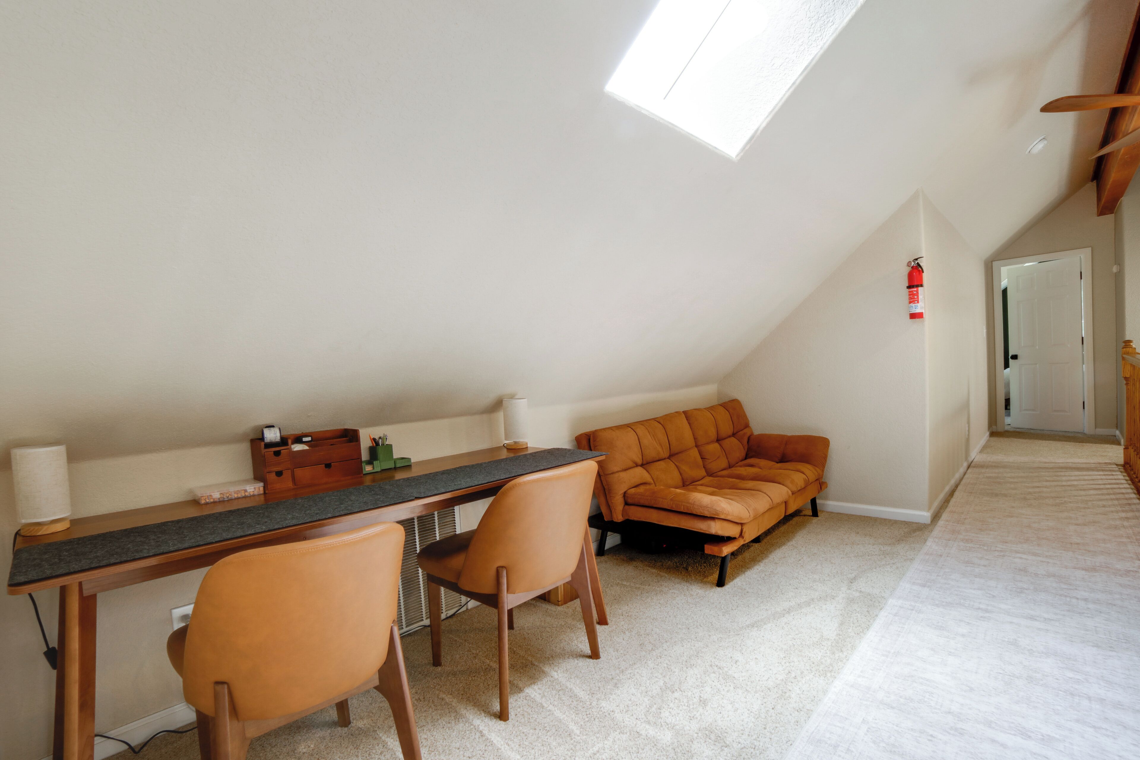 A room with a sloped ceiling, a desk with chairs, a sofa, and a fire extinguisher on the wall.