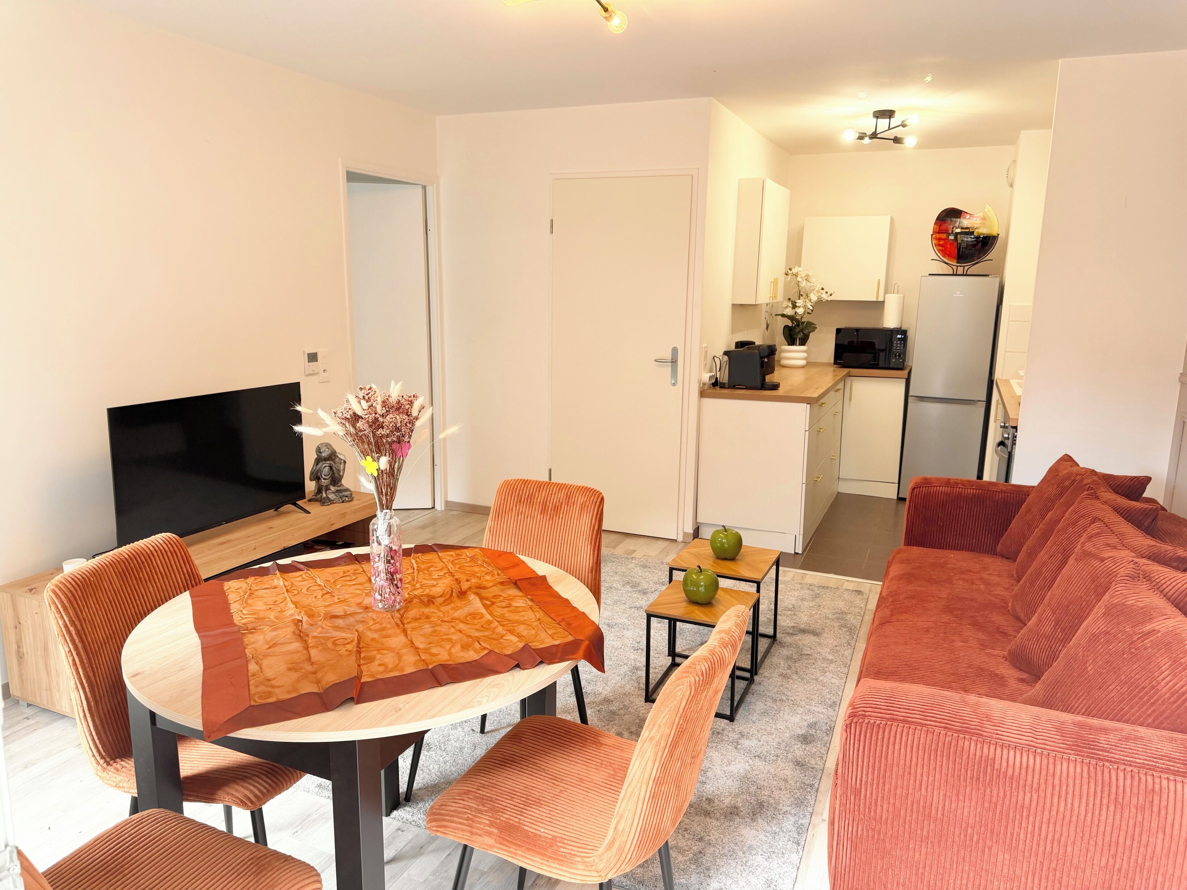 A modern living room with a round wooden table, orange upholstered chairs, a red sofa, a flat-screen TV, and a kitchen in the background.