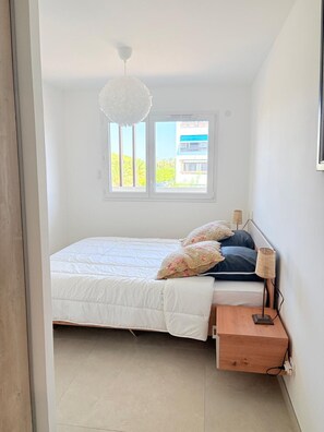 Un dormitorio con cama, mesita de noche, lámpara y ventana con vistas a edificios y árboles.