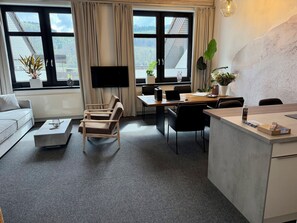 Ein modernes Wohnzimmer mit einer Couch, einem Couchtisch, einem Fernseher, einem Essbereich mit Stühlen, einer Küchenarbeitsplatte und großen Fenstern mit Vorhängen.