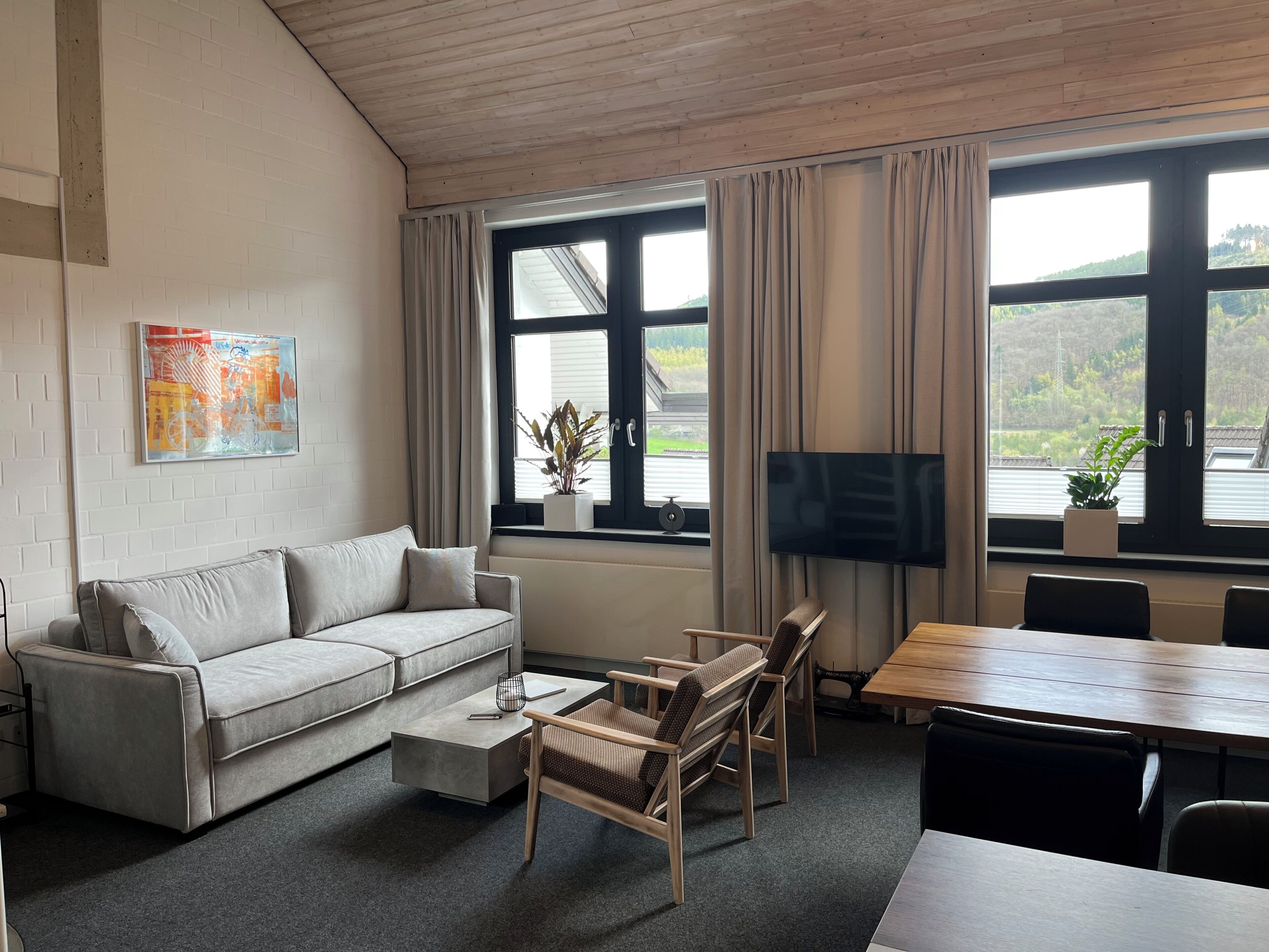 Ein modernes Wohnzimmer mit einer Couch, einem Couchtisch, Sesseln, einem Fernseher und großen Fenstern mit Vorhängen.