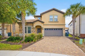 A two-story house with a brick driveway, a garage, and a front yard with palm trees and shrubs. - Windsor Paradise | 8BR Resort Villa Near Disney (Kissimmee)