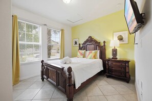 A bedroom with a bed, a nightstand, a lamp, a TV, and a window with blinds. - Windsor Paradise | 8BR Resort Villa Near Disney (Kissimmee)
