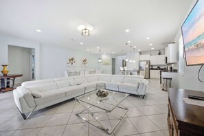 A modern living room with a sectional sofa, a glass coffee table, and a kitchen area in the background. - Windsor Paradise | 8BR Resort Villa Near Disney (Kissimmee)