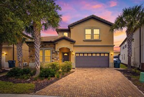 A two-story house with a brick driveway, a garage, and a well-maintained front yard with palm trees and shrubs. - Windsor Paradise | 8BR Resort Villa Near Disney (Kissimmee)