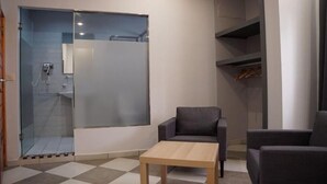 Quarto com box de vidro, mesa de madeira, duas poltronas e um armário com peças penduradas.