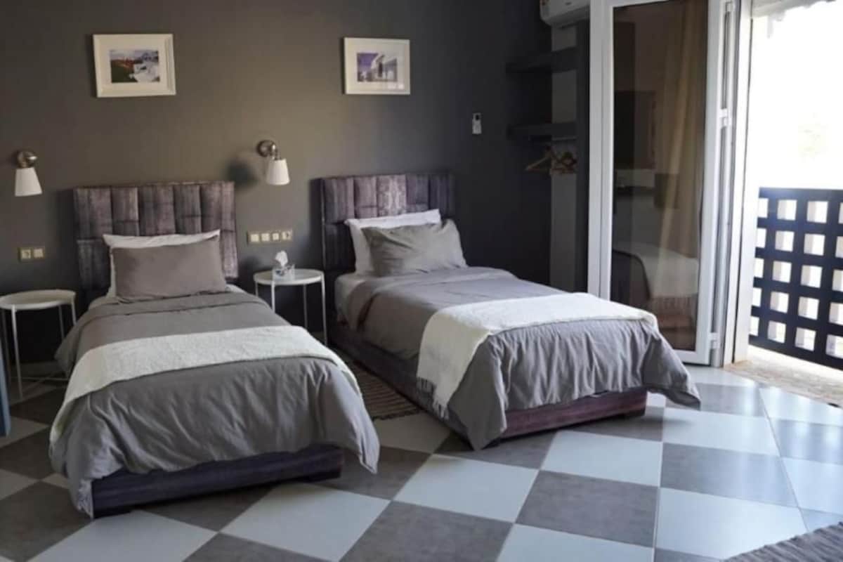 A hotel room with two beds, a small table, a chair, and a view of the outside.