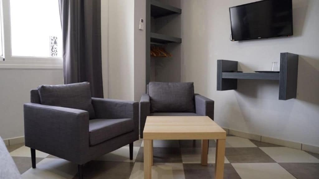 A modern living room with two grey armchairs, a wooden coffee table, a flat-screen TV mounted on the wall, and a built-in shelving unit.