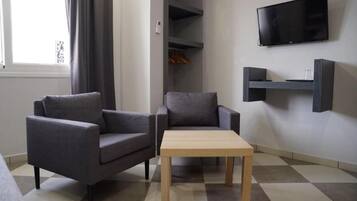 A modern living room with two grey armchairs, a wooden coffee table, a flat-screen TV mounted on the wall, and a built-in shelving unit.