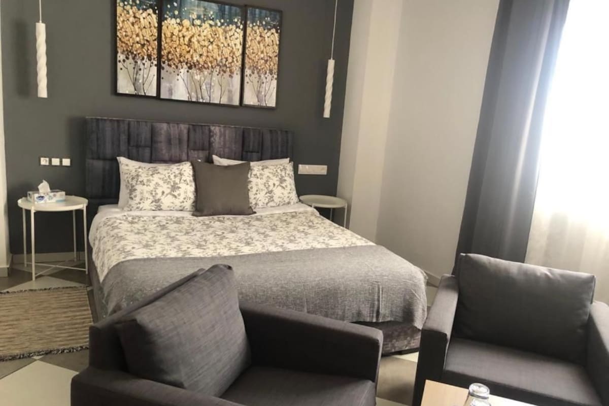 A modern hotel room with a large bed, two bedside tables, a sofa, and a large window.