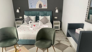 Quarto moderno com uma cama, duas cadeiras verdes, uma mesa de cabeceira com um objeto decorativo e um criado-mudo com abajur.