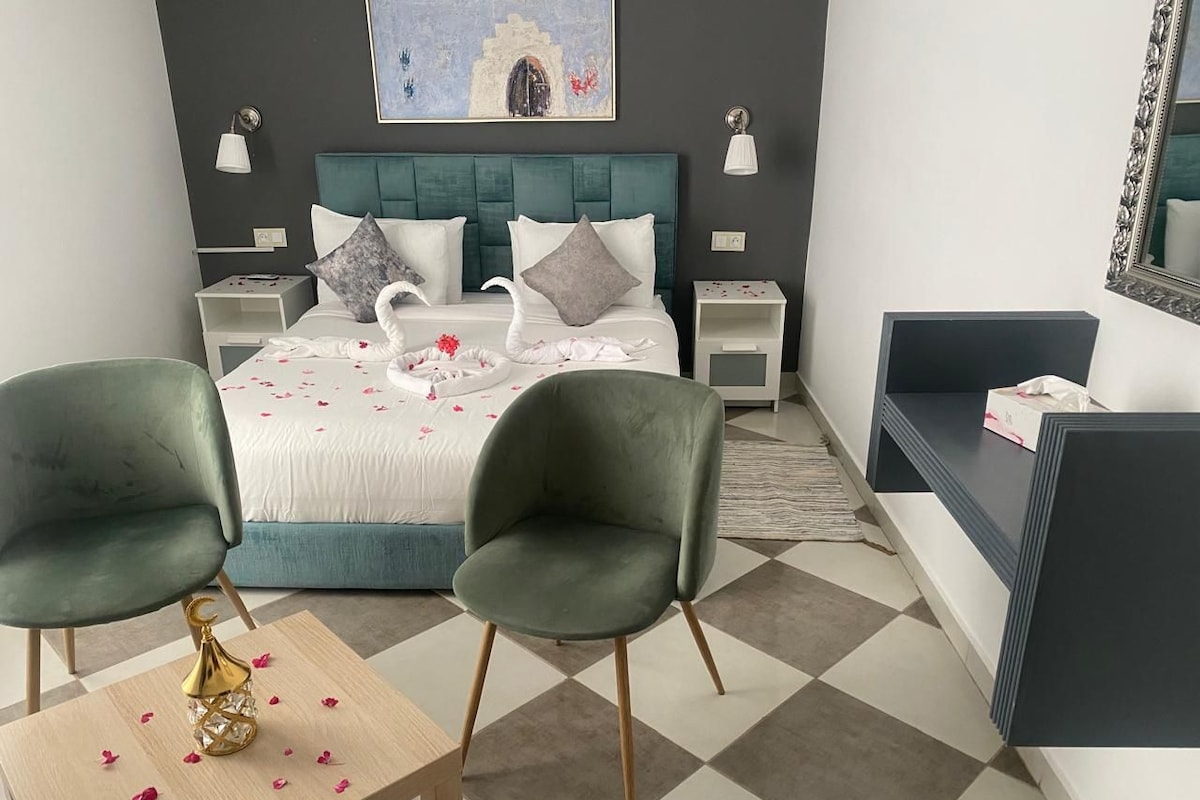 A modern bedroom with a bed, two green chairs, a bedside table with a decorative item, and a nightstand with a lamp.