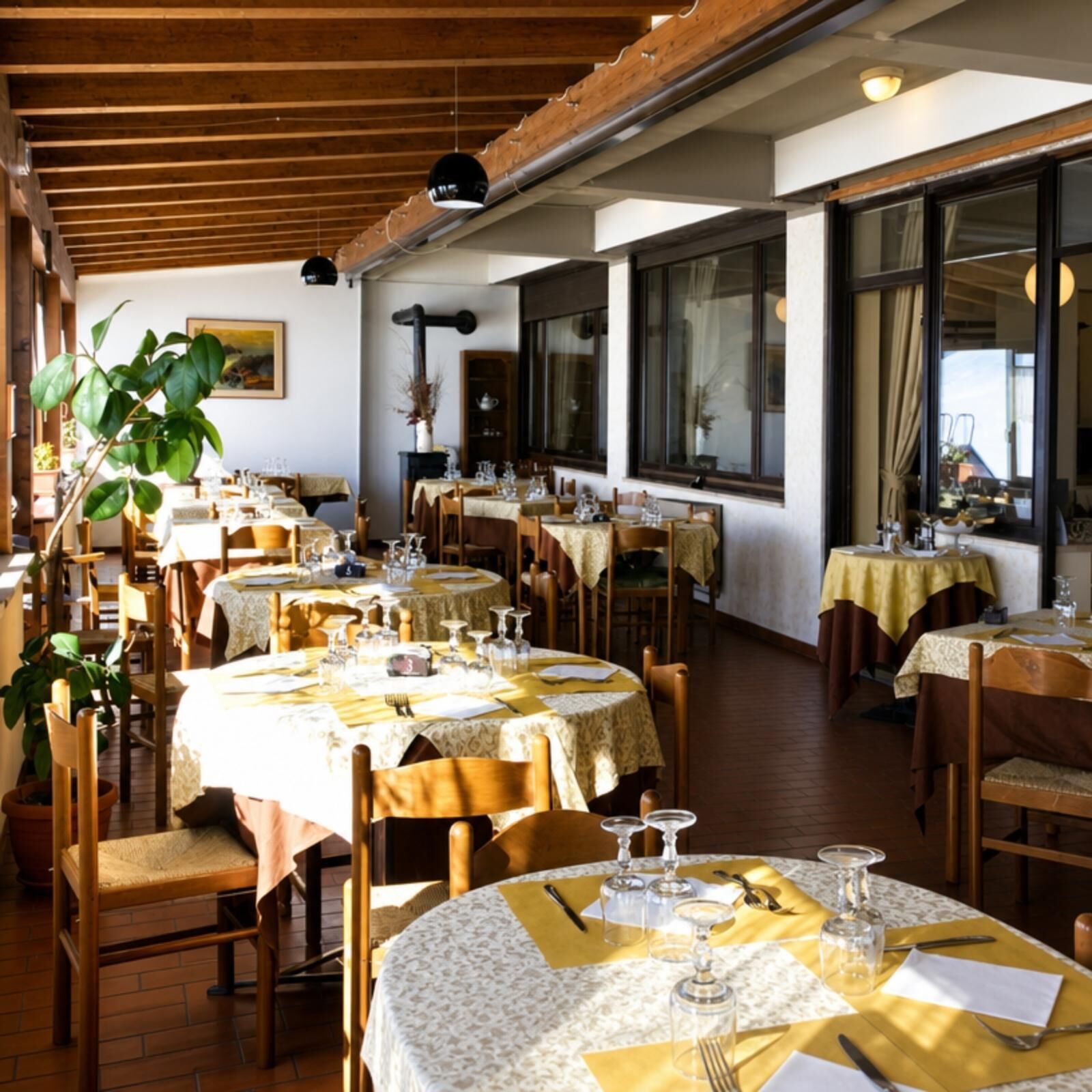 A restaurant with multiple tables set for dining, wooden beams on the ceiling, and large windows.