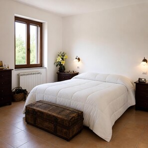A bedroom with a large bed, a wooden chest, a window with a view, and a nightstand with a lamp.