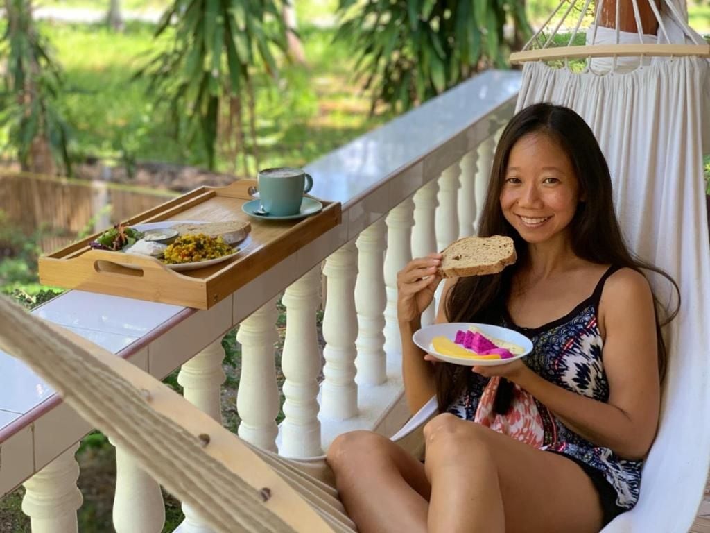 A person seated on a white cushioned chair with a tray containing food and a cup, outdoors with greenery in the background.