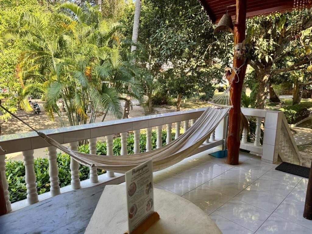 A hammock on a balcony with a view of a garden and a building in the background.