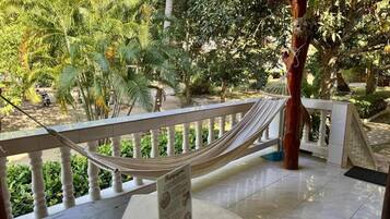 A hammock on a balcony with a view of a garden and a building in the background.