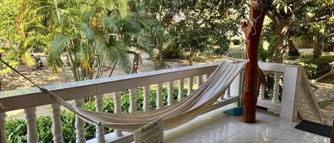 A hammock on a balcony with a view of a garden and a building in the background.