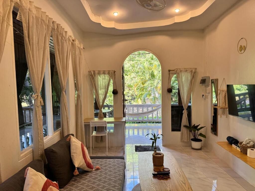 A spacious living area with a sofa, a coffee table, and a TV. There is a balcony with a view of greenery.