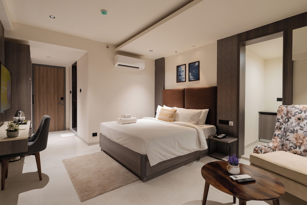 A modern hotel room with a bed, bedside table, chair, desk, and a large mirror.