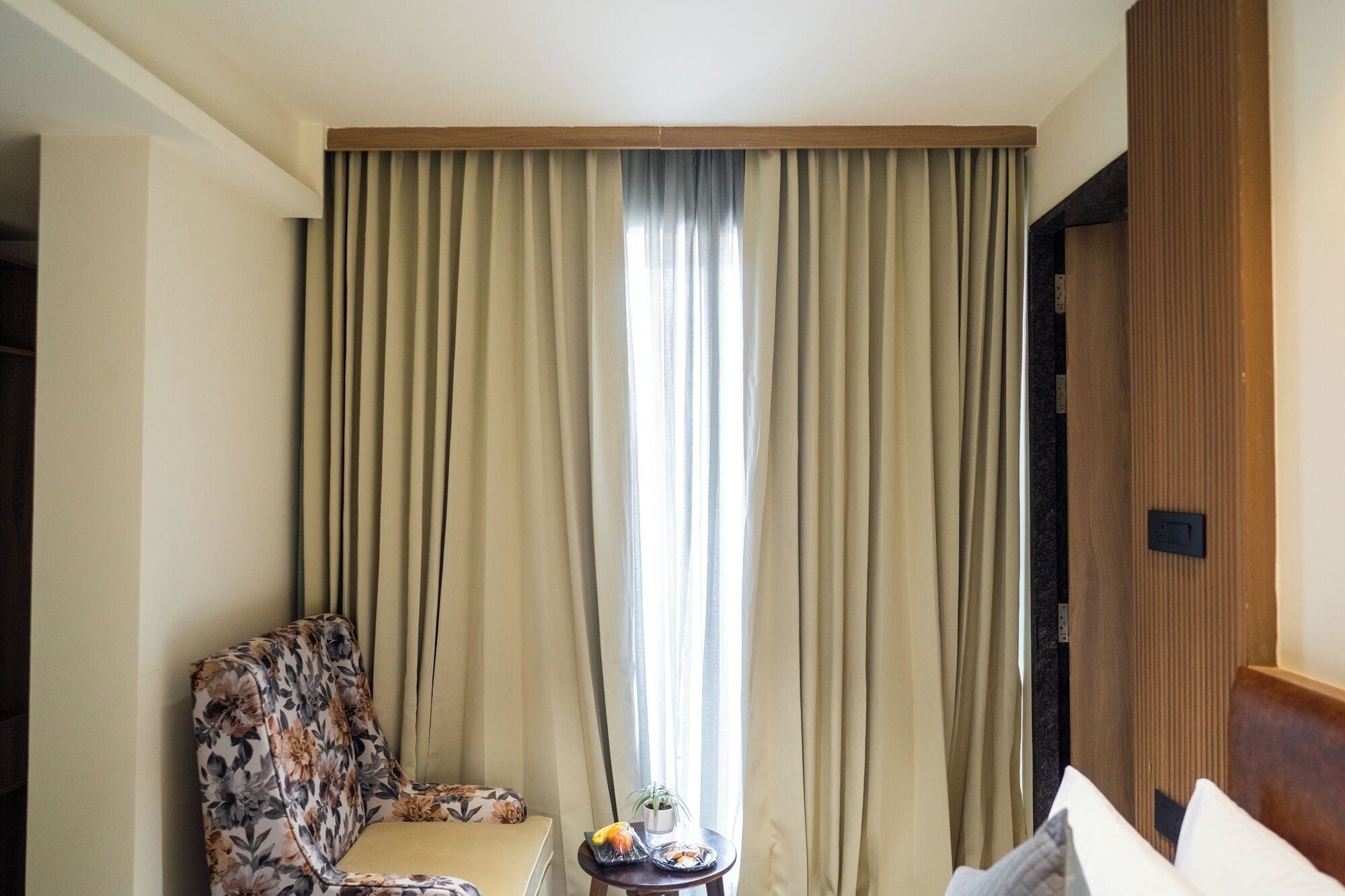 Habitación de hotel con sofá, una mesita con una planta y fruta, y un ventanal con cortinas.