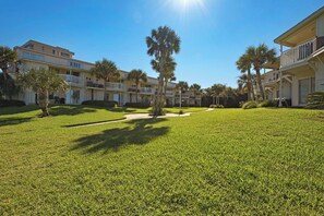 A hotel courtyard with a well-maintained lawn, palm trees, and a clear blue sky. - 🌊🏖️ Sand Dollar W-210 | Steps to Beach + Bunk Room | Pensacola Beach (Pensacola Beach)