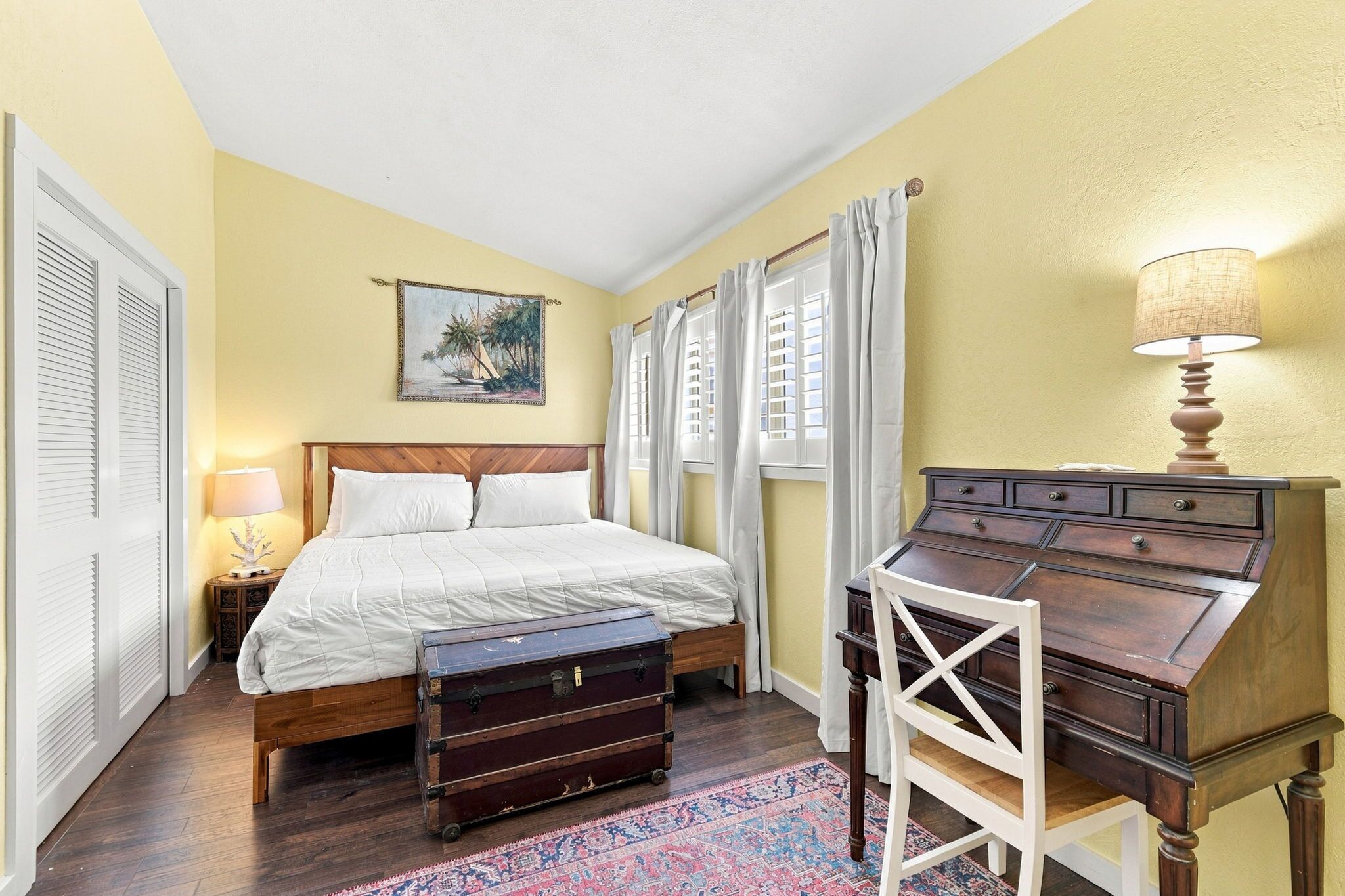 A bedroom with a bed, a dresser, a lamp, a painting, and a window with curtains.