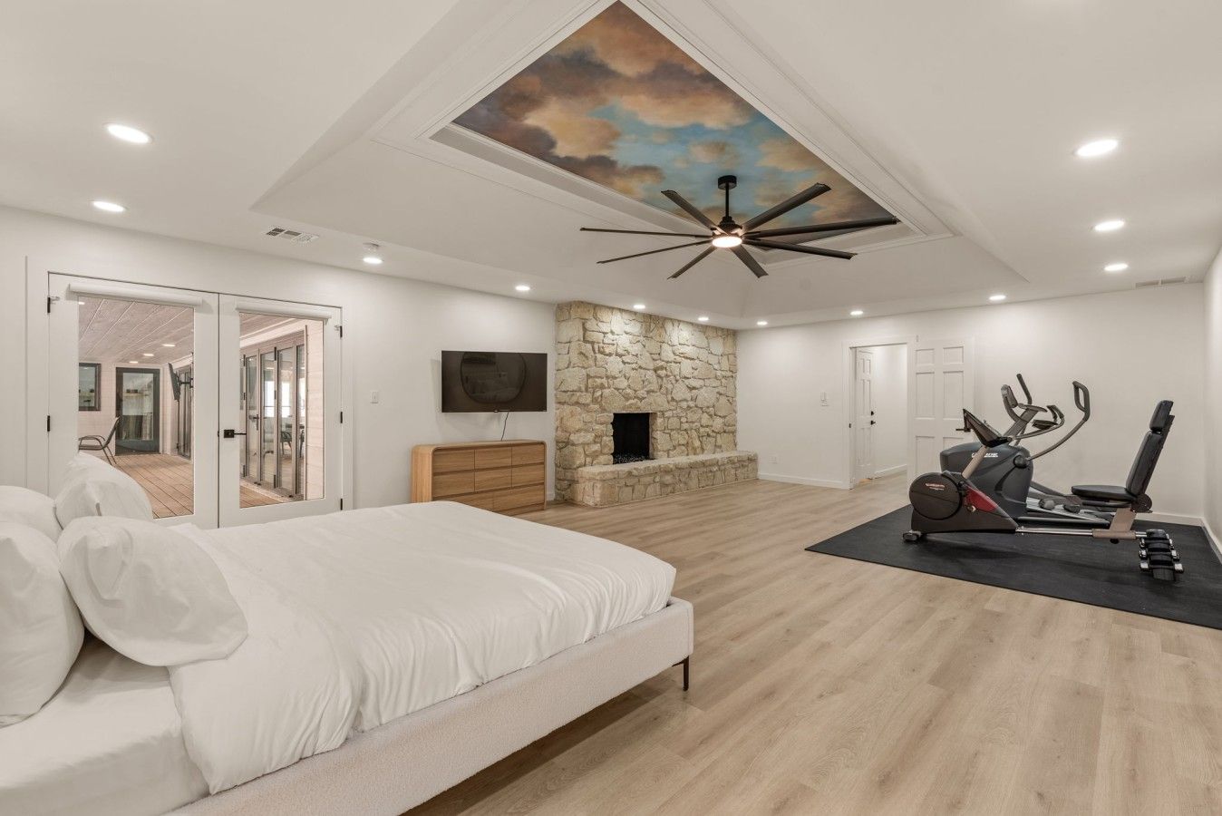 A modern bedroom with a large bed, a stone fireplace, a ceiling fan, and exercise equipment.