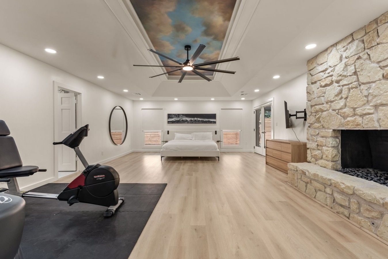 A modern bedroom with a ceiling fan, a stone fireplace, a bed, and a treadmill.