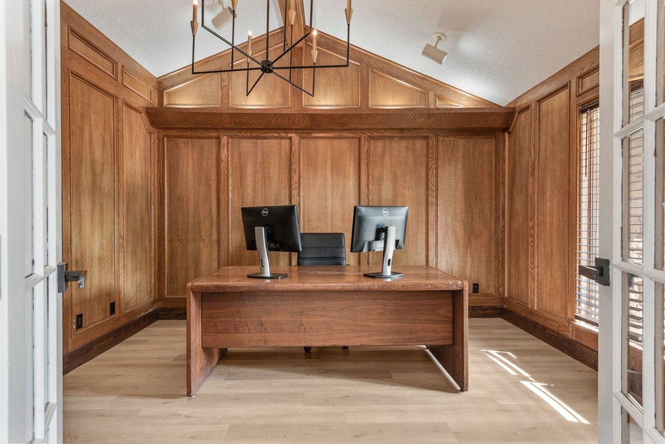 A room with wooden paneled walls, a desk with two monitors, and a modern chandelier.