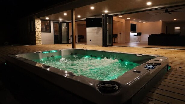 A hot tub with a wooden deck and a modern interior with a seating area. - Luxury South Tulsa Retreat | 4 King Beds • Hot Tub (Tulsa)