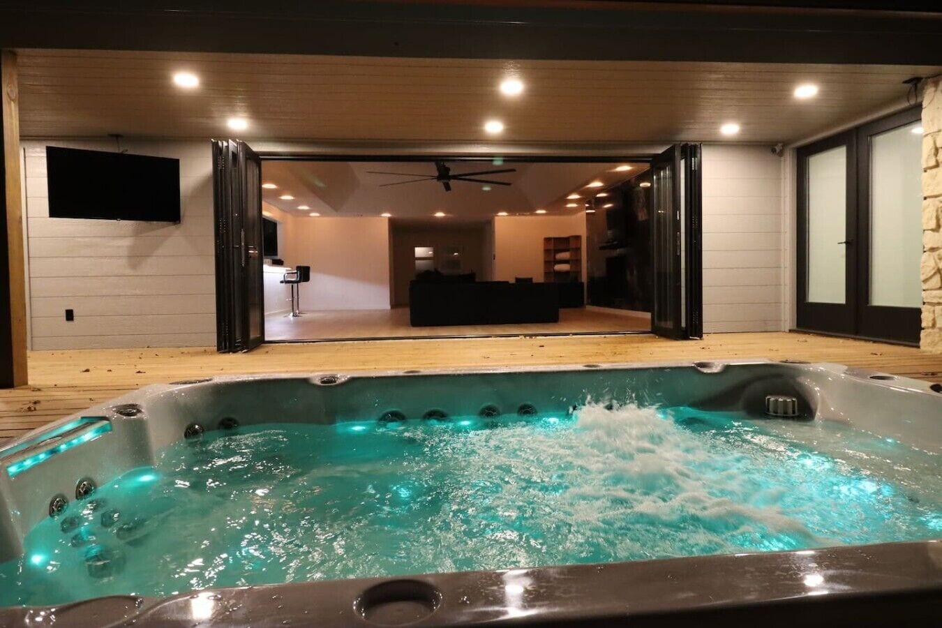 A hot tub with a wooden deck and a view into a modern living area with a ceiling fan.