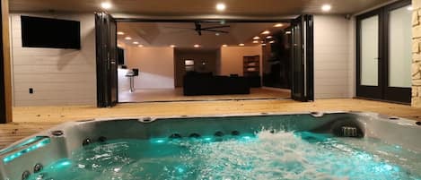 A hot tub with a wooden deck and a view into a modern living area with a ceiling fan.