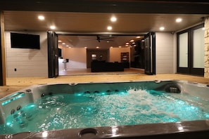 A hot tub with a wooden deck and a view into a modern living area with a ceiling fan. - Luxury South Tulsa Retreat | 4 King Beds • Hot Tub (Tulsa)
